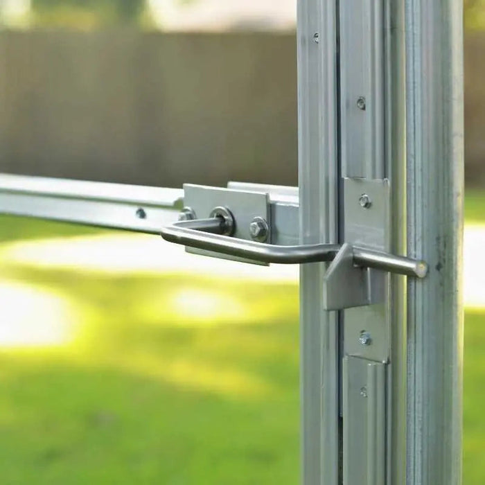 Silver metal greenhouse door latch securing a clear panel frame. Green garden background visible.