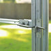 Silver metal greenhouse door latch securing a clear panel frame. Green garden background visible.