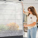 Woman zipping clear, reinforced greenhouse cover on a dark raised garden bed. Ideal for protecting plants, vegetables, and flowers.