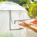 Person's hands opening a white grid greenhouse cover flap, revealing green plants. Great for garden protection & ventilation.