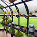 Greenhouse interior with black shelves displaying potted plants, colorful flowers, and gardening tools. Wooden floor, green lawn visible.
