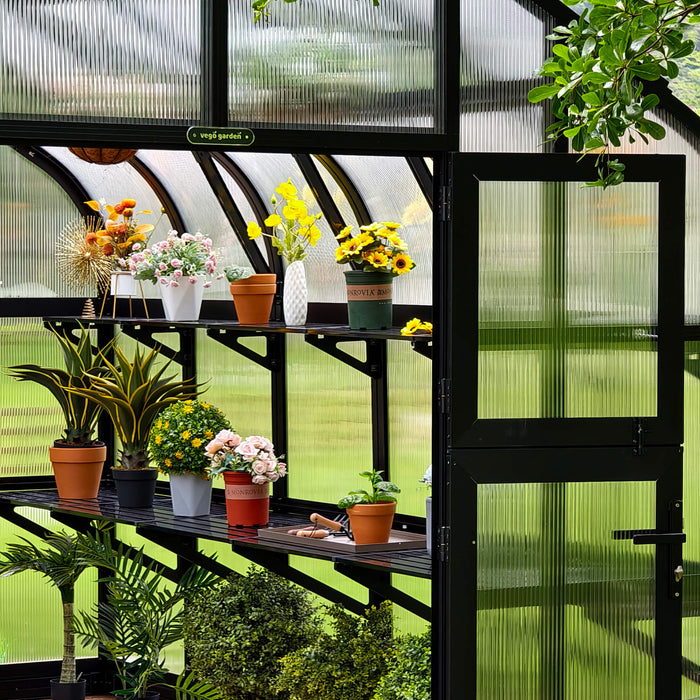 Vego Garden greenhouse interior with black frame, ribbed polycarbonate panels, shelves of potted plants, sunflowers, and gardening tools.