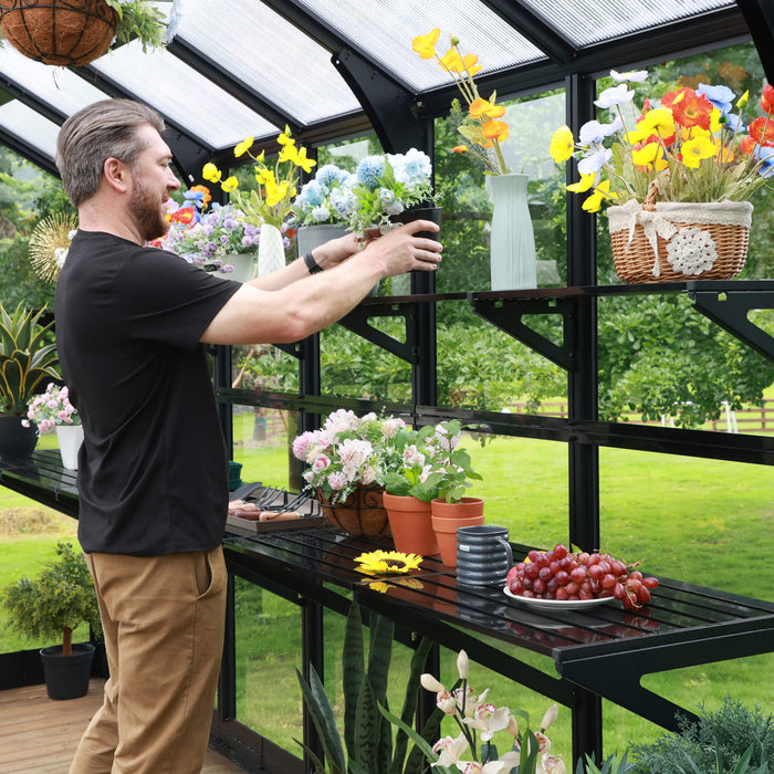 Man arranging vibrant flowers & plants in a modern black greenhouse. Features clear panels, adjustable shelves, patio deck, garden view.