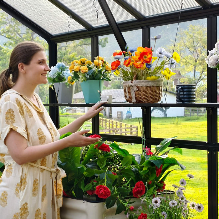 Woman in cream dress admiring red roses, colorful artificial flowers in bright greenhouse with lush garden view.