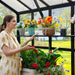 Woman in cream dress admiring red roses, colorful artificial flowers in bright greenhouse with lush garden view.
