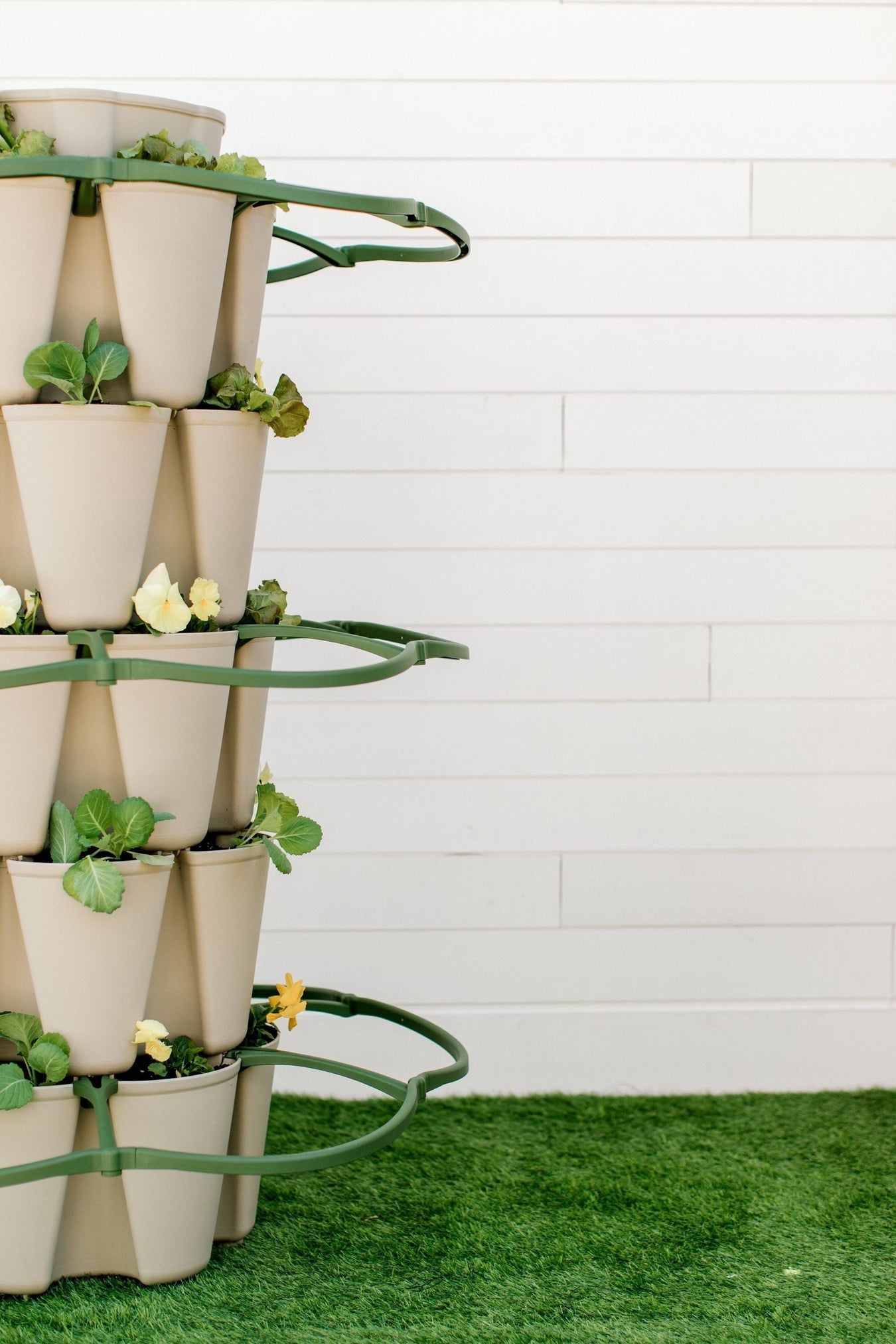 Beige vertical garden tower with fresh lettuce, kale, and yellow pansies on green artificial turf against white wall.