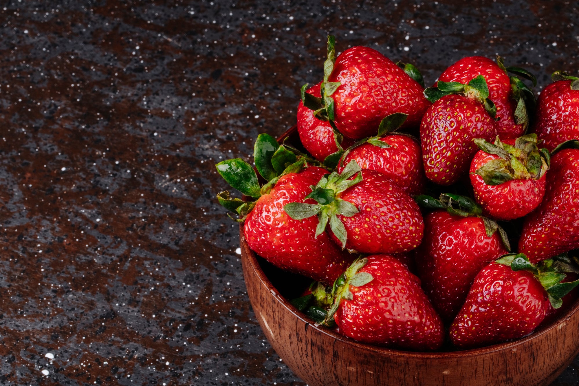 Fresh red strawberries in a wooden bowl on a dark speckled countertop.
