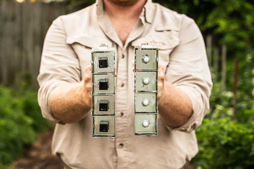 Man holds two modular bee hotels: one with open nesting cavities, one with capped nesting tubes for solitary bees in a garden.